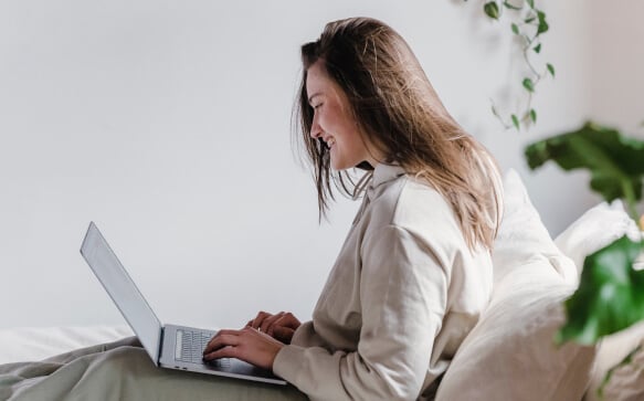 A person sits on a sofa using a laptop, smiling, using Dialogue's EAP program on her laptop. There are green plants in the background.
